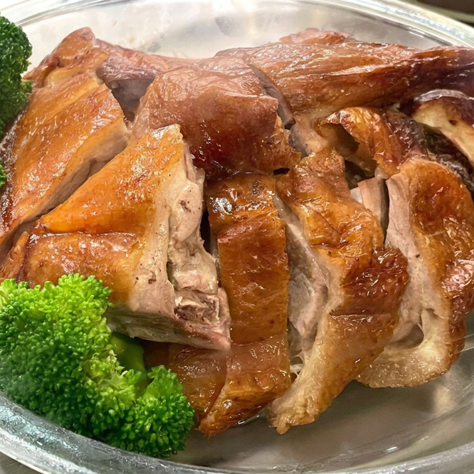 Sliced roasted duck with glossy brown skin served alongside bright green broccoli florets on a clear plate.
